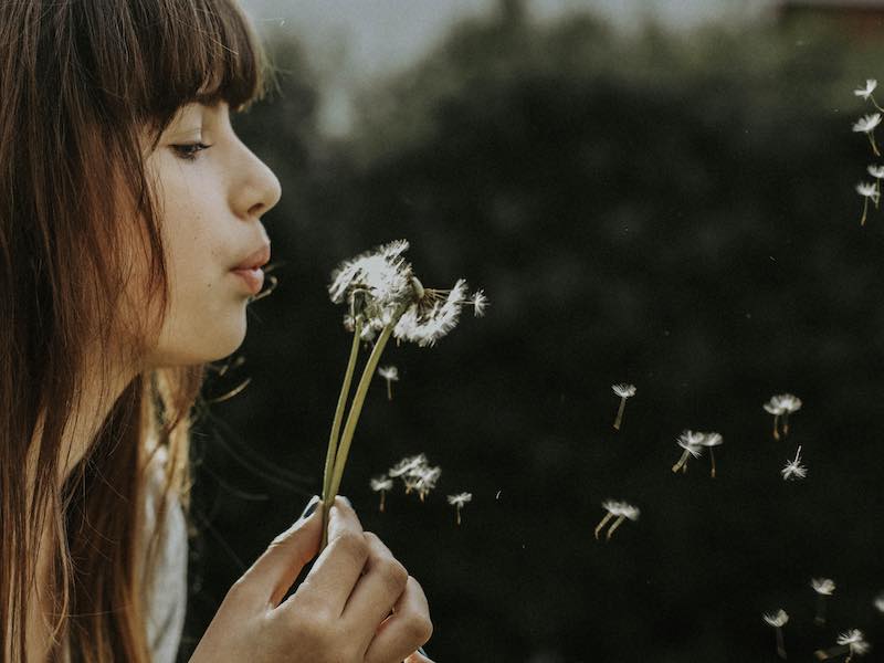 A young woman is in profile against a dark background, gently blowing dandelion seeds.